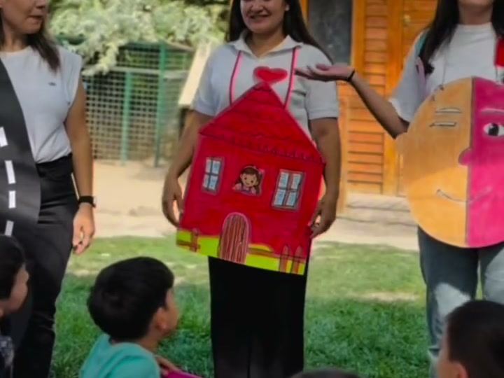 Bahçede elinde kâğıttan ev ve yüz maketleri tutan öğretmenler ve onları izleyen çocuklar